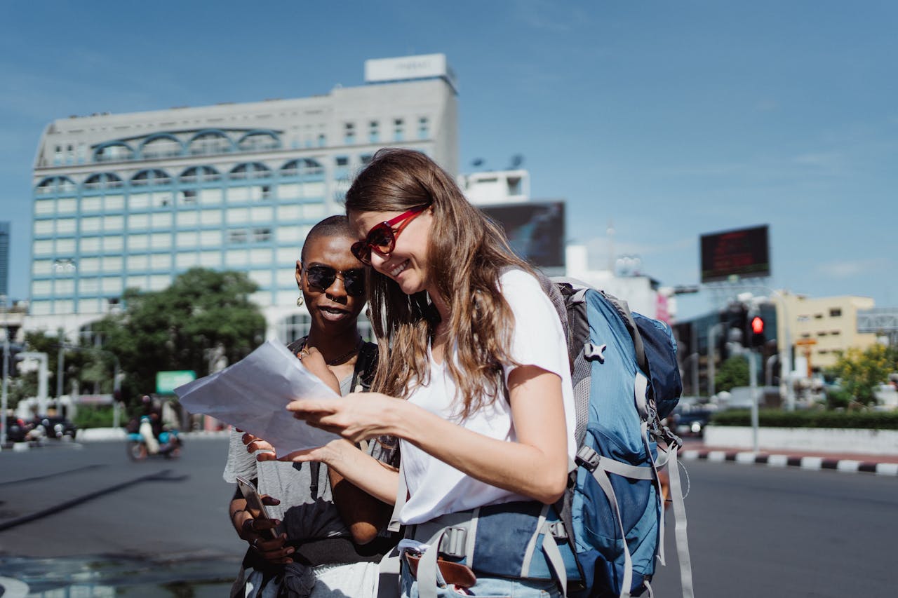 Services-02 Two women tourists with backpacks check a map on a city street under a bright sky.