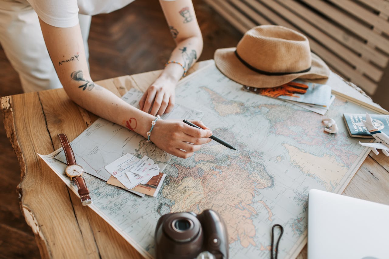 Services-03 Tattooed woman planning a trip with a world map, passports, and travel essentials on a wooden table.